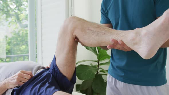 Biracial male therapist examining while lifting senior man's leg at health club