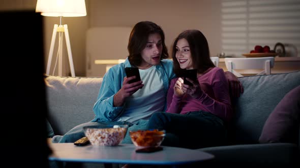 Young Woman Choosing Goods in Her Phone Showing It to Husband and Discussing Choice n Couch at Home