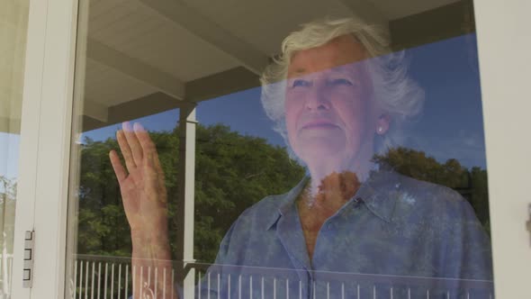 Happy caucasian senior woman at home, looking out of window on sunny day smiling and waving