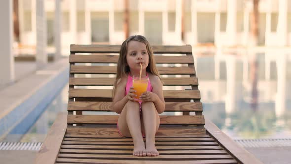 Portrait of Little Cute Girl in Pink Swimsuit with Orange Juice Glass Resting on a Wooden Lounge