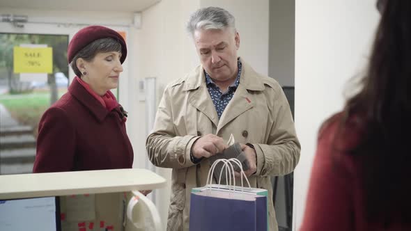 Positive Senior Caucasian Couple Paying for Purchase and Leaving Shop. Unrecognizable Young Woman