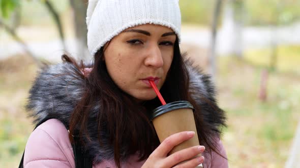 A beautiful woman with long dark hair sits on a park bench and drinks delicious aromatic coffee