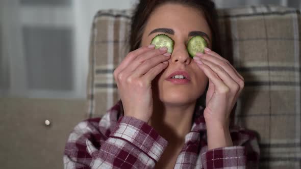 Portrait of Pretty Young Woman in Pajama Making Eye Mask From Cucumbers in Cozy Evening