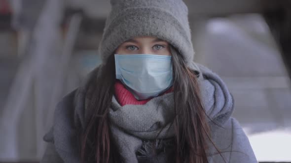 Portrait of Brunette Grey-eyed Girl in Winter Clothes and Protective Mask Looking at Camera. Young