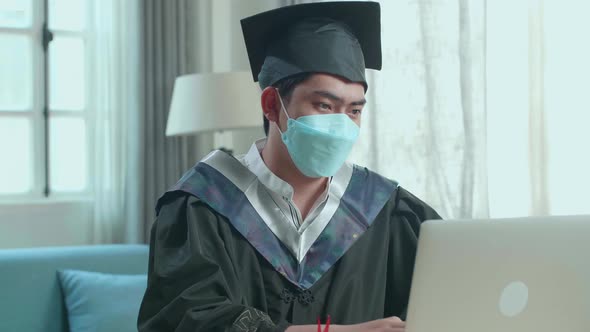 Asian Man Wearing Protection Face Mask And Wearing A Graduation Gown And Cap Use Laptop Computer alt