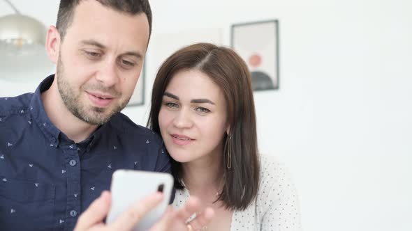 Happy Couple Relaxing on Sofa at Home Husband Holding Smartphone in Hands They Laugh