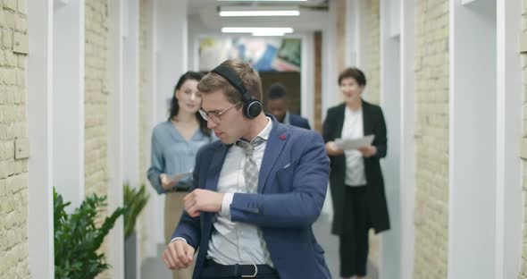 Cheerful Handsome Man in Headphones Dancing in Office Corridor. Joyful Multiethnic Colleagues