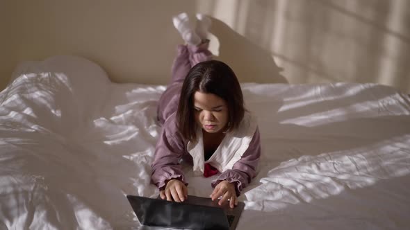 Wide Shot Portrait of Thoughtful Young Little Woman in Pajamas Surfing Internet on Laptop Lying on