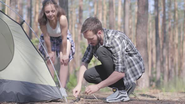 Young Man Putting Up a Tent in a Pine Forest While His Girlfriend Standing Near. Nature Lovers