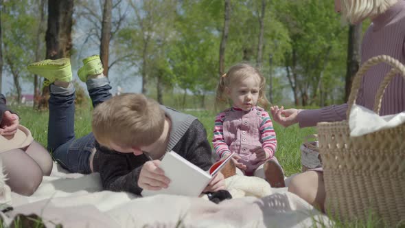 Teenage Blond Boy Lying on the Blanket in the Park Reading the Book, Cute Little Girl in a Pink