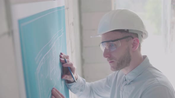 Young engineer-architect in a bright room in a helmet and glasses works with blueprints 