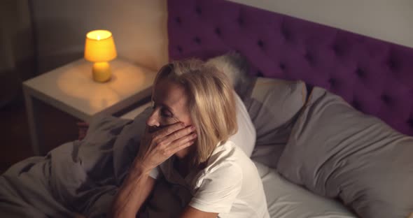 Senior Woman Sitting in Bed and Feeling Stressed with Husband Sleeping on Background