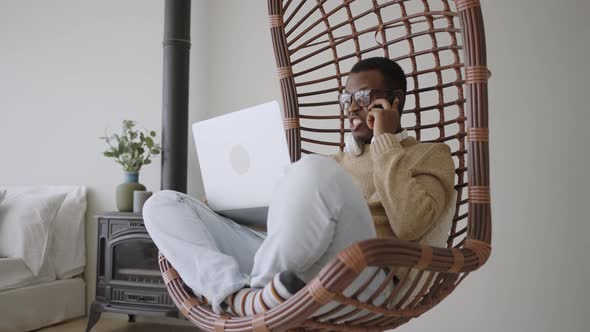 Black Freelancer Talks on Phone Swinging in Basket Armchair