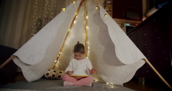 Little Girl on Evening Play and Watch Cartoons on a Tablet in a Tent