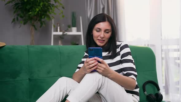 Woman with Dark Hair Sitting on Green Sofa Near Window and Typing Message to Somebody on Her Phone