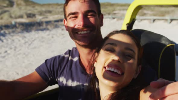 Happy caucasian couple sitting in beach buggy by the sea using smartphone taking selfies