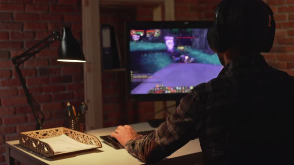 Back View of Caucasian Male Sitting in Headphones Playing Computer Game in Light of Table Lamp