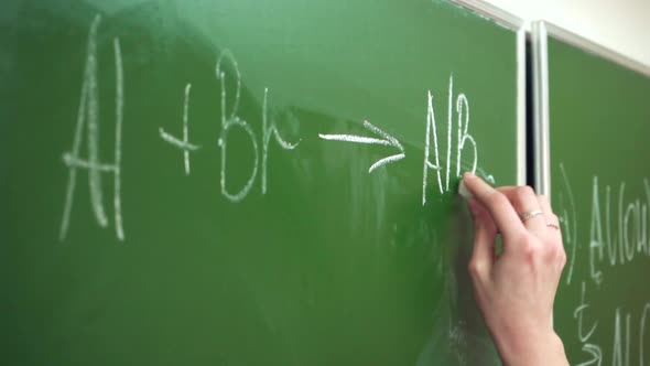 A hand with a piece of chalk draws a chemical formula on a green chalkboard alt