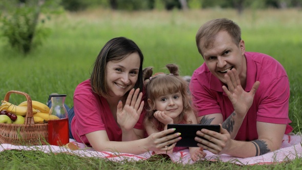 Family Weekend Picnic. Daughter Child Girl with Mother and Father Play Games on Tablet, Waving Hands
