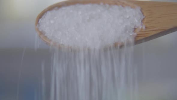 Close-up of White Sugar or Salt Granules Falling Out of Wooden Spoon. Unknown Person Adding