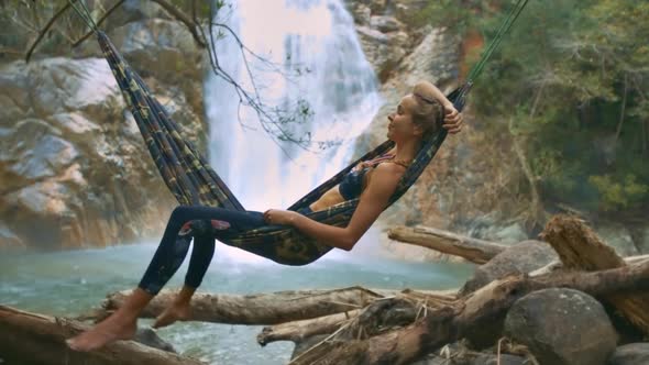 Girl Rests in Hammock Above Trunks Near Waterfall