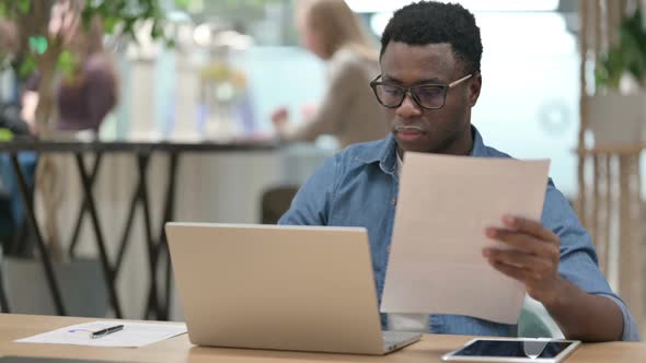 Young African Man Reading Documents While Working on Laptop