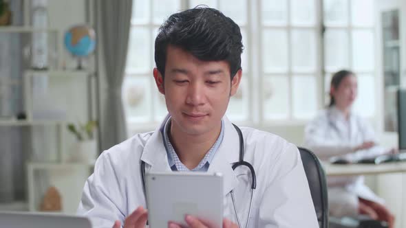 Young Asian Man Doctor Is Using Tablet While Work With Laptop Computer In Workplace