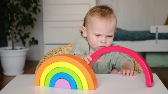 Little Baby Child in Clothes Made of Natural Fabric Plays with Rainbow Colored Wooden Toys at Table