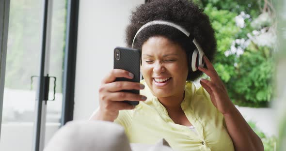 Video of happy plus size african american woman with headphones on sofa, listening to music