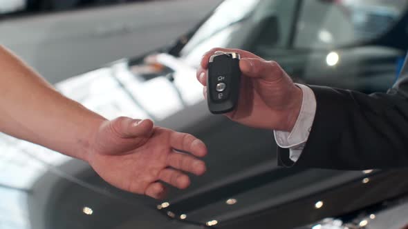 Young Man Giving Keys of Car To Buyer. Men Shaking Hands in Beautiful Car Dealership on Background