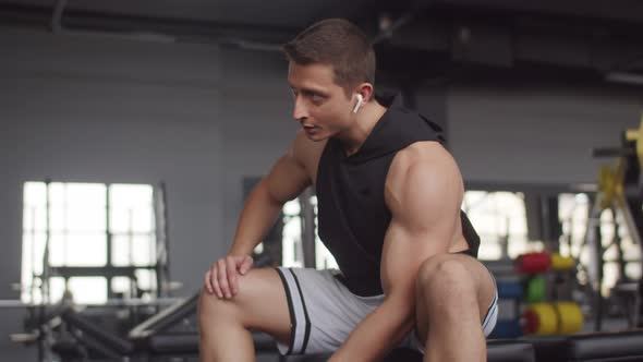 Man with Headphones is Sitting in Gym and Lifting Dumbbell with One Hand Front View