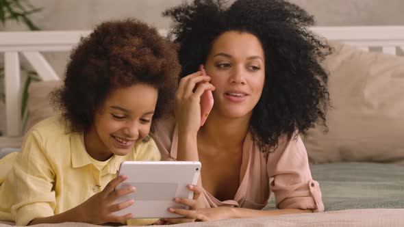 African American Woman is Talking on Smartphone Litle Girl is Playing on Tablet