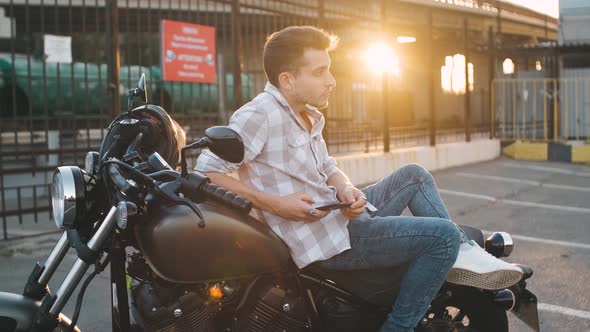 Young Man Motorcyclist with Smart Phone in Hands on Custom Motorcycle on Street