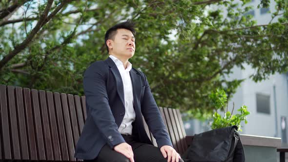 Male asian businessman relax, meditates outdoors sitting on a bench on urban