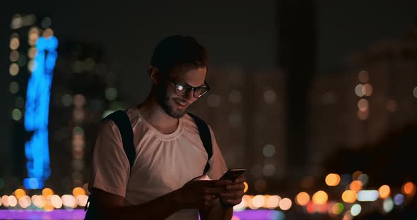 A Young Man with Glasses at Night Looks Into the Screen of a Smartphone