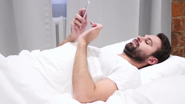 Beard Man in Bed Browsing on Smartphone at Night