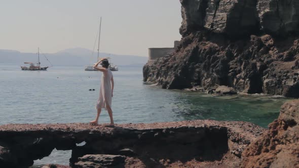 A woman walks along a rocky beach watching the small boats and catamaran. Inside the Santorini calde