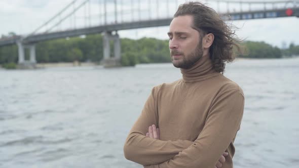 Depressed Bearded Brunette Man with Curly Hair Standing on River Bank and Holding Head in Hands