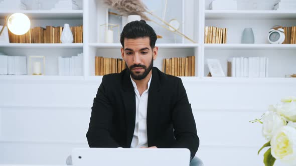 Hispanic Businessman in a Suit Sitting By Desk in Front of Laptop Working From Home in Online