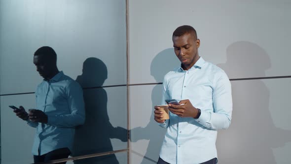 Afro American Man Is Drinking Coffee in Street at Morning, Viewing News Tape in Smartphone