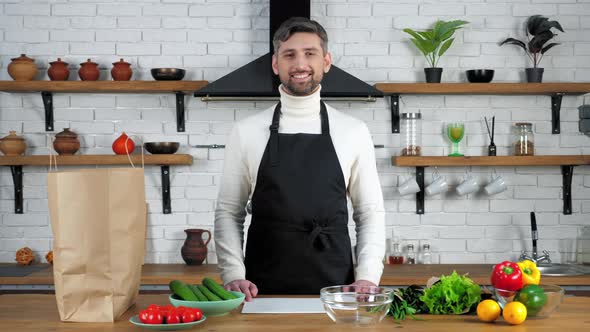 Portrait smiling bearded man in black apron looking camera at home kitchen