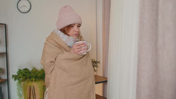 Young Sick Woman Wear Hat Wrapped in Plaid Stay Alone Shivering From Cold on Sofa Drinking Hot Tea