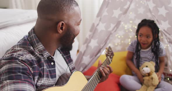 Happy african american father and daughter sitting in blanket tent, playing guitar, clapping hands