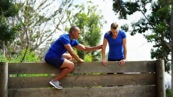 Male trainer assisting fit woman to climb over wooden wall during obstacle course 4k