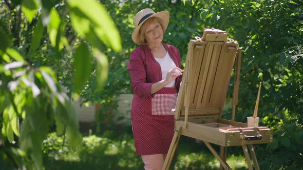 Portrait of Talented Senior Caucasian Woman Painting in Slow Motion on Easel Standing in Sunny