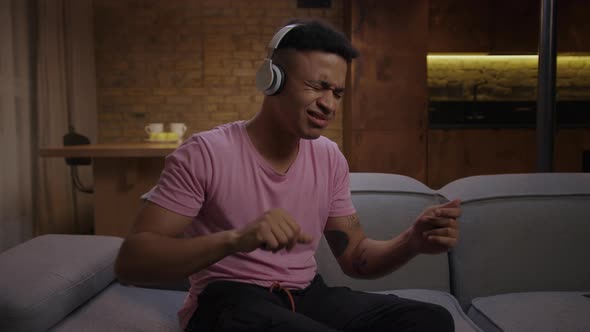 Young African American Man Enjoys Listening To Music in White Wireless Headphones Sitting on Couch