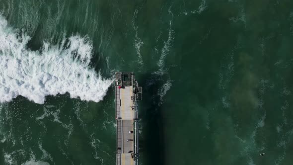 Large waves crashing under a concrete walking platform with people sightseeing . High drone view loo