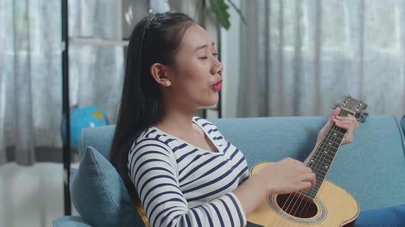 Close Up Of Asian Woman Relaxing Leaning On Sofa To Sing And Play A Guitar At Home
