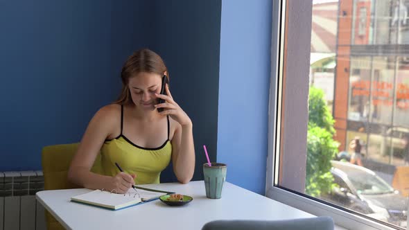 A Young Female Freelancer Sits in a Cafe Talking on a Mobile Phone and Writing Diary Entries