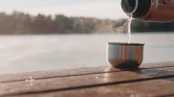 Man Pouring Hot Drink From Thermos in a Cup on a Lake Dock on a Winter Sunny Day